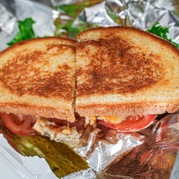 Grilled sandwich with chicken, tomato, and lettuce, served with a pickle spear in foil.