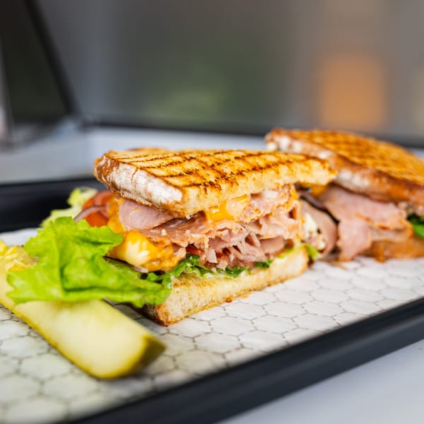 A grilled deli sandwich with meat, cheese, lettuce, and tomato, served with a pickle spear.