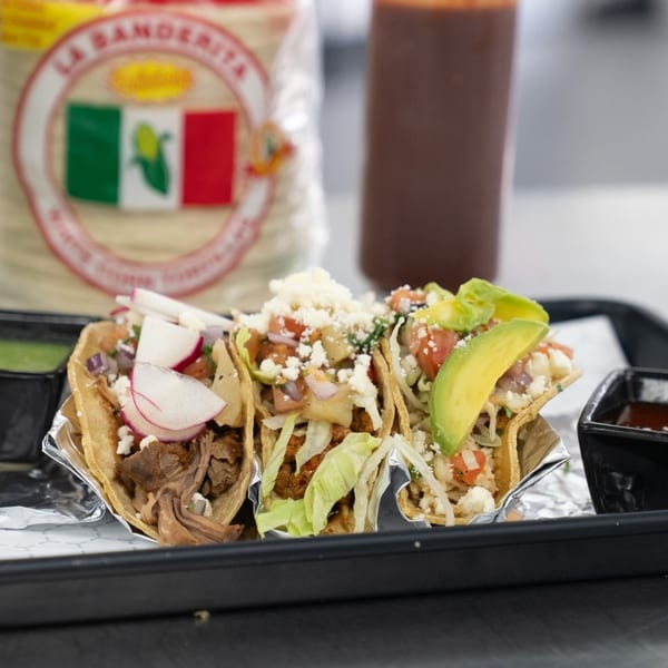 Three deli tacos with various toppings, sauces, and a tortilla package on a black tray.