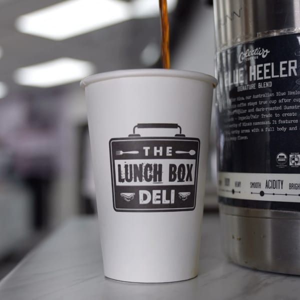 Coffee pouring into a white paper cup with The Lunch Box Deli logo, next to a coffee dispenser.