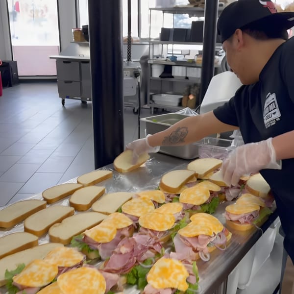 A deli worker wearing gloves assembling many ham and cheese sandwiches on a metal counter.