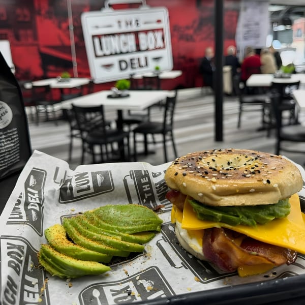 A bagel sandwich with bacon, cheese, and avocado, served with sliced avocado at The Lunch Box Deli.